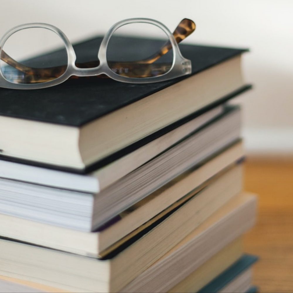 A stack of books sitting on a table. A pair of glasses rest on the top of the pile.