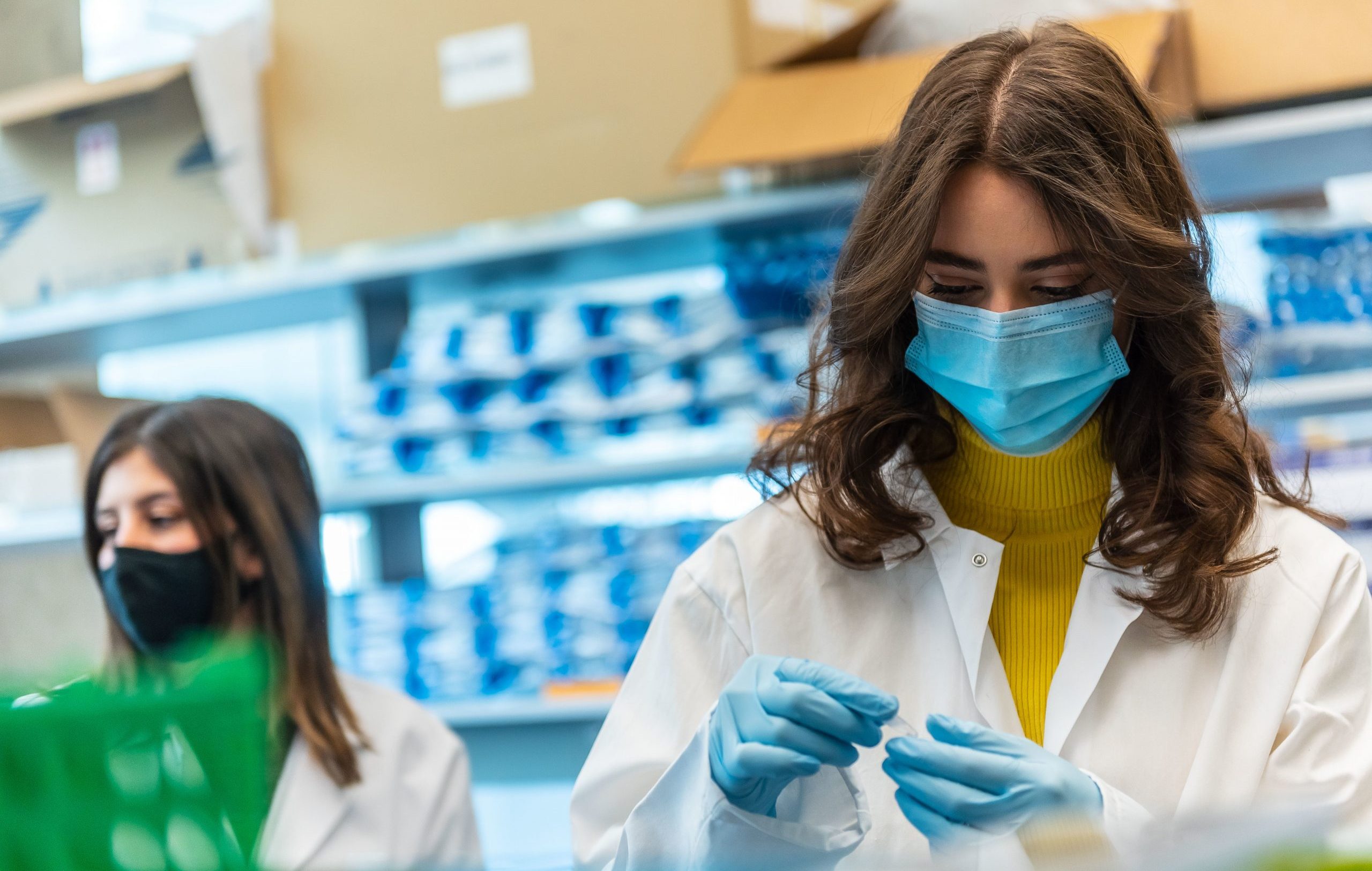 A laboratory. In the foreground, a student in a white lab coat, blue mask, and blue gloves holds a small plastic vial. In the background, another student in a black mask is in front of a shelf filled with supplies.