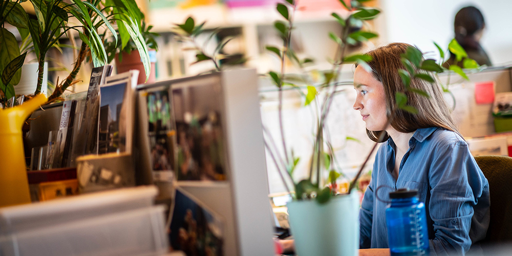 A woman works on a computer in an office filled with plants and personal touches.