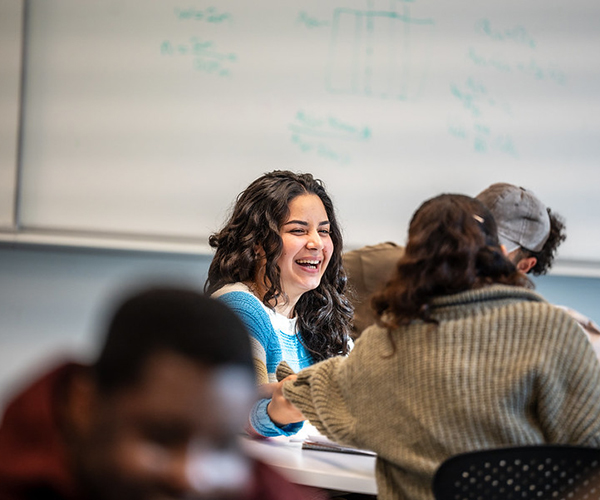 Three students sit laughing at a desk with a whiteboard behind them. 